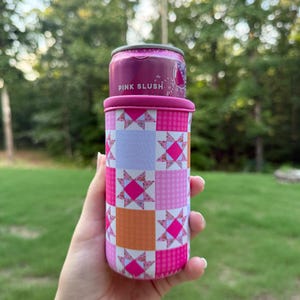 Puede incluir: Un enfriador de latas rosa y naranja con un patr&oacute;n de patchwork de cuadrados con motivos de estrellas y dise&ntilde;os a cuadros. El enfriador sostiene una lata de metal con una etiqueta rosa que muestra claramente el texto PINK SLUSH en letras blancas.