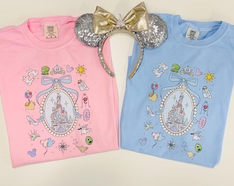 May include: Two t-shirts, one pink and one light blue, with a castle graphic and various cartoon icons. A sequined headband with mouse ears and a gold bow sits between the shirts. The shirts are folded and ready to wear.