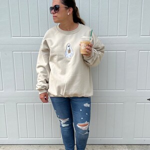 May include: A woman wearing a light beige sweatshirt with a ghost holding a coffee cup graphic. The sweatshirt has a crew neck and long sleeves. She is wearing blue jeans with rips in the knees and is barefoot. She is holding a cup of coffee in her right hand.