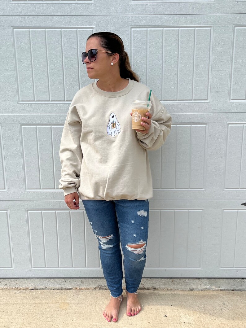 May include: A woman wearing a light beige sweatshirt with a ghost holding a coffee cup graphic. The sweatshirt has a crew neck and long sleeves. She is wearing blue jeans with rips in the knees and is barefoot. She is holding a cup of coffee in her right hand.