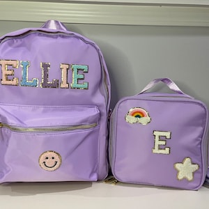 May include: Purple backpack and lunch bag set with personalized patches. The backpack has the name "ELLIE" on the front and a smiley face patch. The lunch bag has a rainbow patch, the letter "E" and a star patch.
