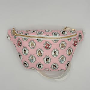 May include: Pink fanny pack featuring a repeating pattern of framed princess portraits. The bag has a gold zipper and a white adjustable strap. The design includes images of various Disney princesses.