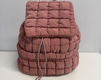 May include: A quilted pink backpack with a drawstring closure. The backpack has a square quilted pattern.