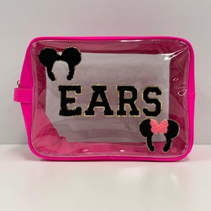 May include: A bright pink, rectangular cosmetic bag with a clear front. The word "EARS" is spelled out in black, gold-trimmed letters. Black Mickey Mouse ear appliqués are on the bag, one with a pink bow. A zipper runs along the top.
