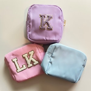 May include: Three small zippered pouches with different colored fabric and embroidered letters. The pouches are purple, pink, and light blue. The purple pouch has a gold glitter letter "K" embroidered on it. The pink pouch has gold glitter letters "LK" embroidered on it. The light blue pouch has no embroidery.
