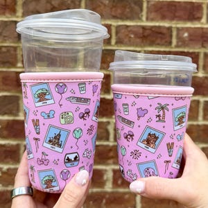 May include: Two clear plastic cups with light pink neoprene sleeves featuring a repeating pattern of colorful illustrations including polaroid-style photos, monorails, palm trees, churros, balloons, and mouse-ear headbands on a light pink background.