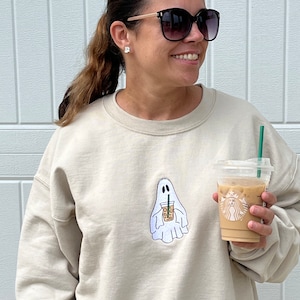 May include: A beige sweatshirt with a white ghost holding a coffee cup with a green straw. The ghost has a small face with two eyes and a mouth. The coffee cup has a brown lid and a white logo with a mermaid on it.