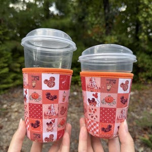 May include: Two clear plastic cups with orange-trimmed fabric sleeves featuring a patchwork pattern of red and pink squares. The design includes illustrations of a castle, pumpkins, teddy bears, and mouse-ear icons with a fall-themed aesthetic.