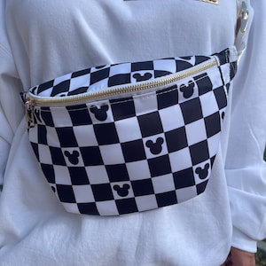 May include: A black and white checkered fanny pack with gold-colored zipper and Mickey Mouse silhouettes. The fanny pack is worn over a white sweatshirt with the word "HOME" in pink and gold lettering.