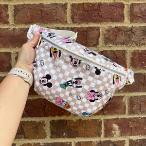 May include: A beige and white checkered fanny pack featuring cartoon characters like Mickey Mouse, Minnie Mouse, and Goofy. The bag has a gold zipper and a white strap. The bag is held in front of a brick wall.