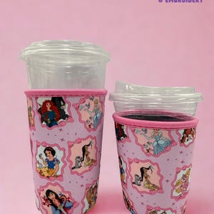 May include: Two pink drink sleeves with clear plastic lids. The sleeves feature a repeating pattern of Disney princesses in pink frames. The top and bottom edges of the sleeves are trimmed in pink. The text "Honey & Embroidery" is in the upper right corner.