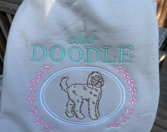 May include: Beige crewneck sweatshirt with embroidered text and graphic. The words "club DOODLE" are in teal, with a pink floral wreath around a doodle dog illustration. The text "DITZY SINCE '85" is in teal.