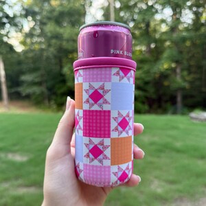 Puede incluir: Una funda de neopreno para latas con un patr&oacute;n de patchwork repetitivo en tonos rosa, naranja y morado claro. El dise&ntilde;o incluye cuadrados alternos de colores s&oacute;lidos, cuadros vichy y motivos de estrellas florales. La parte superior es rosa.