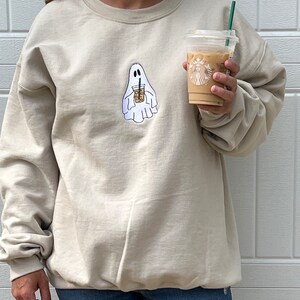 May include: A beige sweatshirt with a ghost holding a coffee cup graphic. The ghost is white and the coffee cup is brown with a white lid and a green straw. The sweatshirt is a crew neck style.