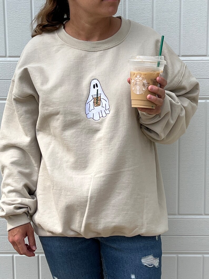 May include: A beige sweatshirt with a ghost holding a coffee cup graphic. The ghost is white and the coffee cup is brown with a white lid and a green straw. The sweatshirt is a crew neck style.