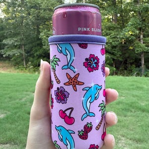 May include: A light purple neoprene can cooler sleeve featuring a repeating pattern of blue dolphins, orange starfish, pink hibiscus flowers, red cherries, strawberries, and palm trees. The sleeve holds a pink can labeled PINK SLUSH.