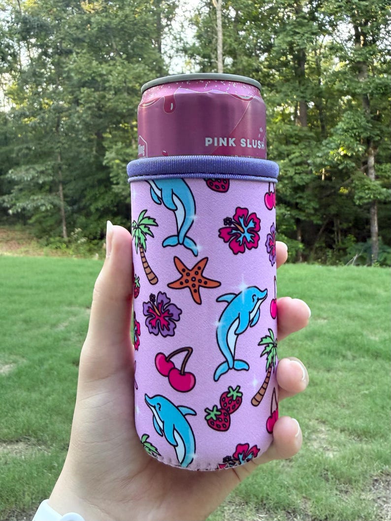 May include: A light purple neoprene can cooler sleeve featuring a repeating pattern of blue dolphins, orange starfish, pink hibiscus flowers, red cherries, strawberries, and palm trees. The sleeve holds a pink can labeled PINK SLUSH.