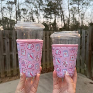 May include: Two clear plastic cups with lids, each fitted with a pink fabric sleeve. The sleeves are decorated with a repeating pattern of framed illustrations, bows, and dresses. The cups are held up against a blurred outdoor background.