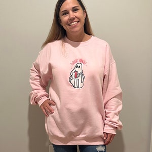 May include: A pink sweatshirt with a white ghost holding a red cup and the text "basic ghoul" printed on the front.