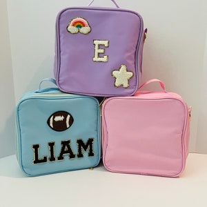 May include: Three lunch bags with personalized patches. The top bag is purple with a rainbow, a star, and the letter "E". The middle bag is blue with a football and the name "LIAM". The bottom bag is pink.