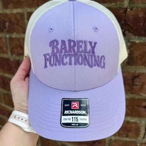 May include: A light purple trucker hat with white mesh back panels. The front features the embroidered text BARELY FUNCTIONING in a darker purple thread. A black rectangular sticker on the brim displays the Richardson 115 Low-Pro Trucker logo.