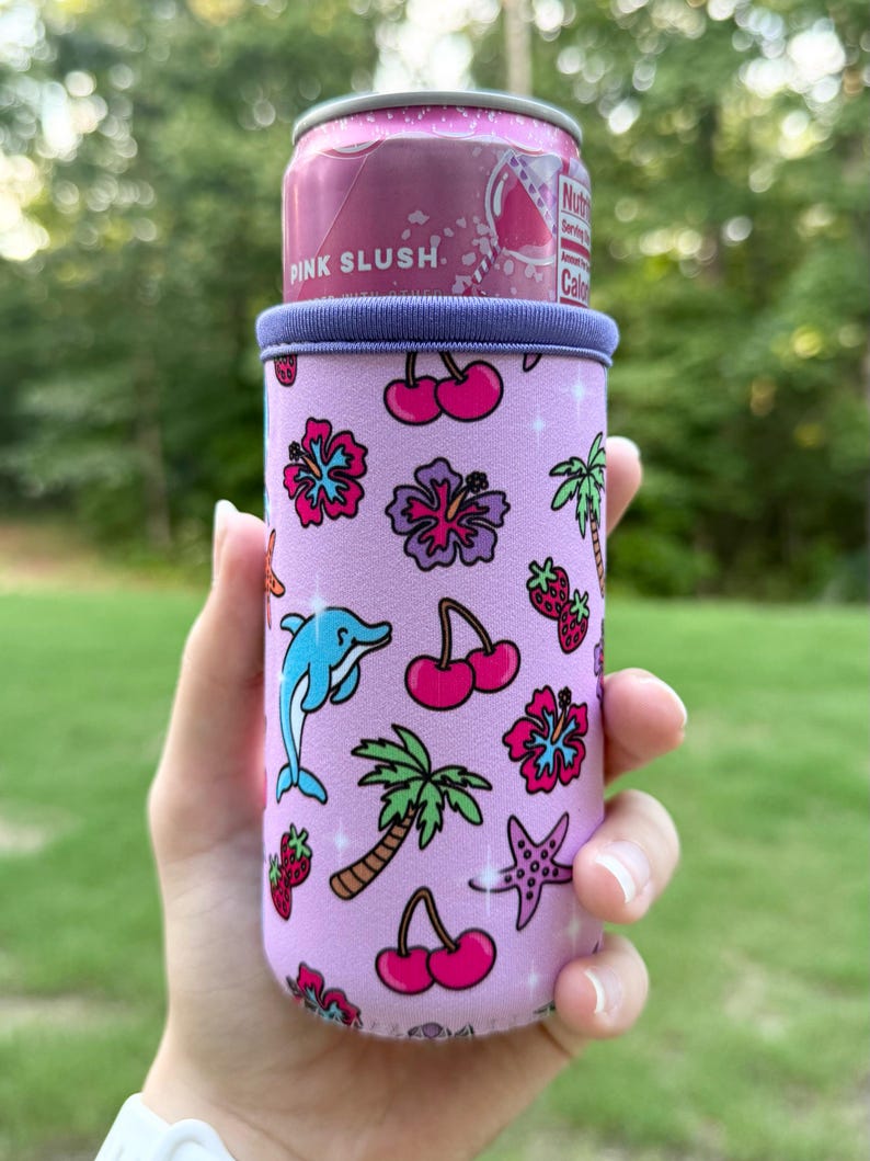 May include: A light purple neoprene can cooler sleeve featuring a repeating pattern of pink hibiscus flowers, red cherries, green palm trees, blue dolphins, purple starfish, and red strawberries, holding a pink slush beverage can.