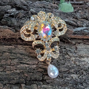 May include: A gold-tone metal brooch featuring an intricate filigree design encrusted with clear rhinestones. The center displays an oval iridescent cabochon, and a single white teardrop-shaped faux pearl hangs from the bottom of the piece.