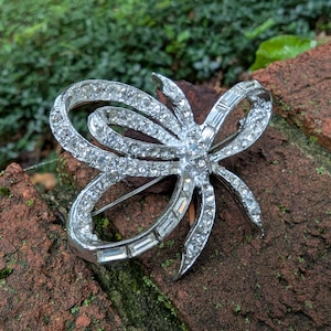 May include: A silver-toned metal brooch shaped like a stylized bow, featuring a combination of round pavé-set clear rhinestones and rectangular baguette-cut clear crystals arranged along the curved loops and trailing ends of the design.