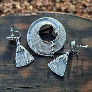 May include: A silver-toned jewelry set featuring a circular brooch and matching clip-on earrings. The brooch has a spiral design with a textured mesh pattern. The earrings have a triangular shape with a tassel-like detail. The set is displayed on a dark wooden surface.