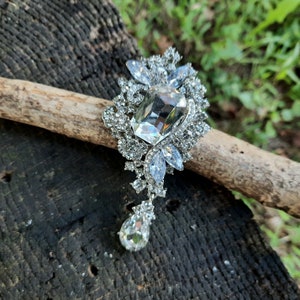 May include: A silver-toned brooch with a large, clear rectangular center stone and numerous smaller, clear crystals. The brooch has a floral design with a dangling teardrop-shaped crystal. The brooch is displayed on a piece of wood.