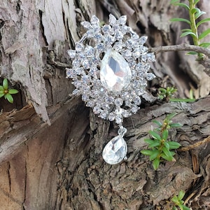 Può includere: Un fermaglio d'argento con un grande ciondolo a goccia di cristallo trasparente circondato da cristalli più piccoli. Il fermaglio è su una superficie di legno marrone ruvida.