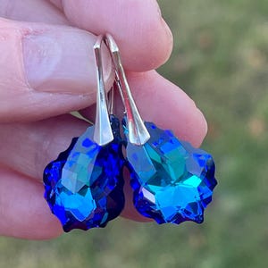 May include: A pair of faceted, teardrop-shaped earrings. The earrings feature a vibrant, deep blue color with hints of teal, and are suspended from silver-colored hooks. The earrings are held against a blurred green background.