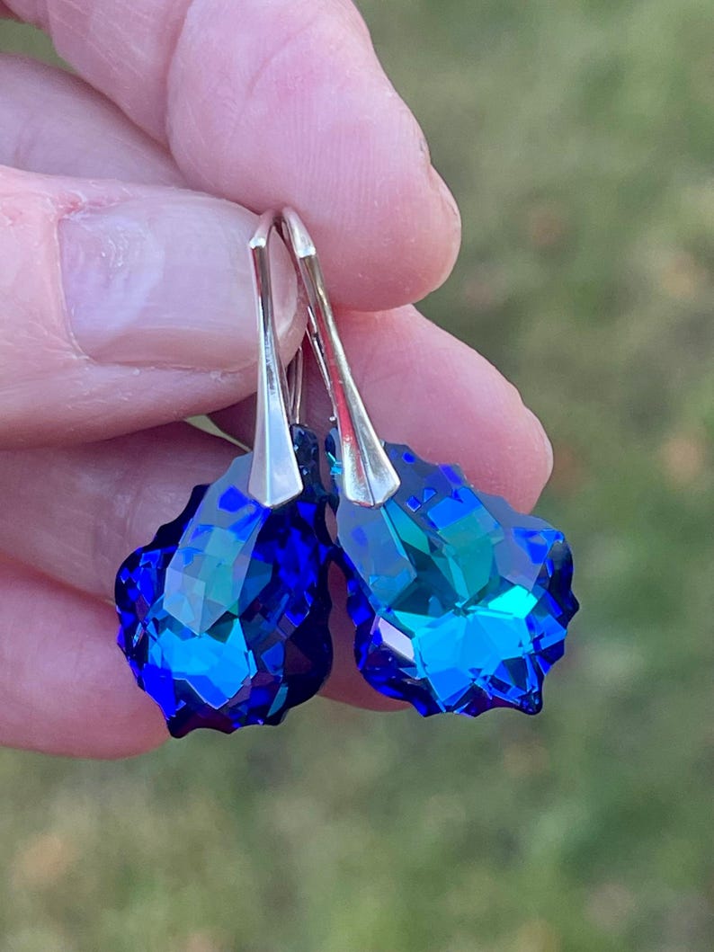 May include: A pair of faceted, teardrop-shaped earrings. The earrings feature a vibrant, deep blue color with hints of teal, and are suspended from silver-colored hooks. The earrings are held against a blurred green background.