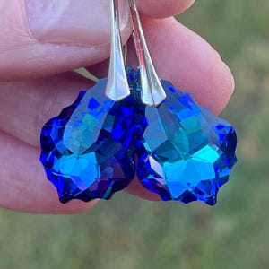 May include: A pair of faceted, teardrop-shaped earrings. The earrings feature a deep blue crystal with a silver-colored hook. The crystals have multiple facets that reflect light, creating a sparkling effect. The earrings are held up against a blurred green background.