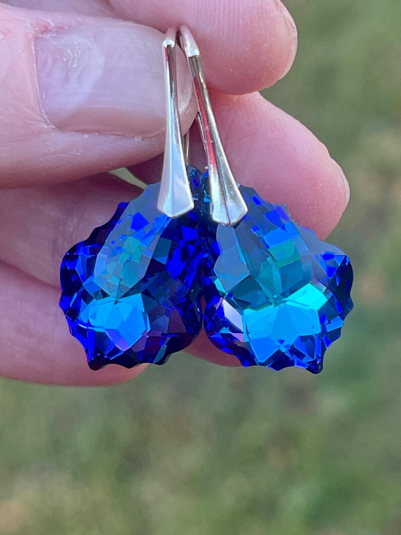 May include: A pair of faceted, teardrop-shaped earrings. The earrings feature a deep blue crystal with a silver-colored hook. The crystals have multiple facets that reflect light, creating a sparkling effect. The earrings are held up against a blurred green background.