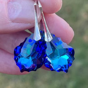 May include: A pair of faceted, deep blue crystal earrings with silver-colored hooks. The earrings have a geometric, multi-faceted design, reflecting light and creating a sparkling effect. The background is blurred, suggesting an outdoor setting.