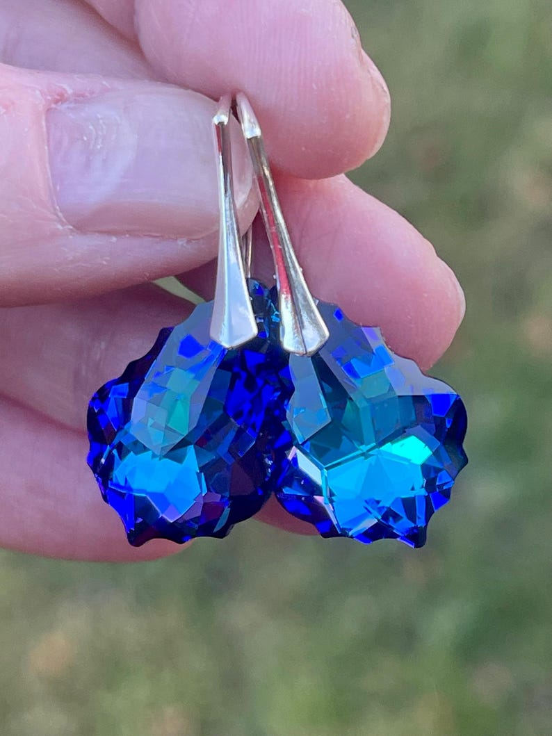 May include: A pair of faceted, deep blue crystal earrings with silver-colored hooks. The earrings have a geometric, multi-faceted design, reflecting light and creating a sparkling effect. The background is blurred, suggesting an outdoor setting.