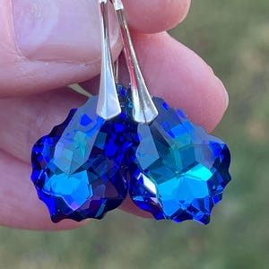 May include: A pair of faceted, deep blue crystal earrings with silver-colored ear wires. The earrings have a heart-like shape and reflect light, showcasing their brilliant color and cut. The background is blurred.