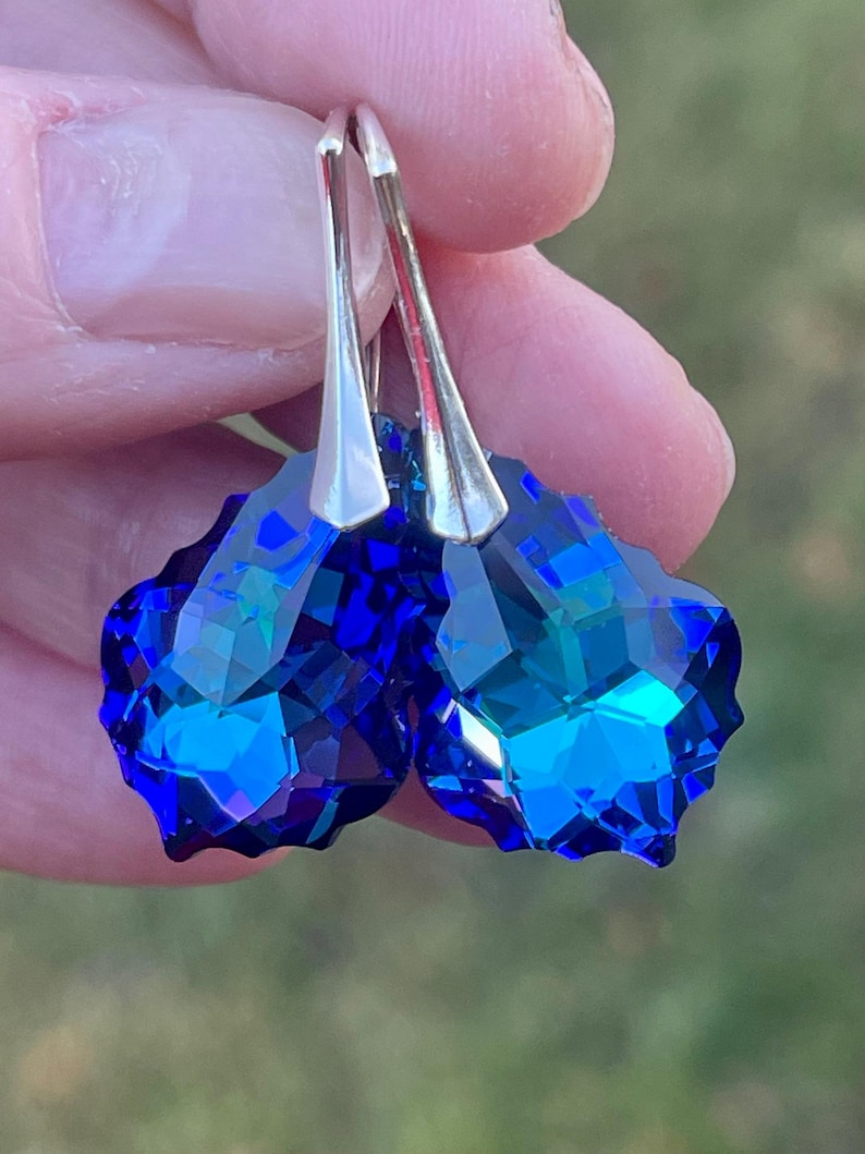 May include: A pair of faceted, deep blue crystal earrings with silver-colored ear wires. The earrings have a heart-like shape and reflect light, showcasing their brilliant color and cut. The background is blurred.