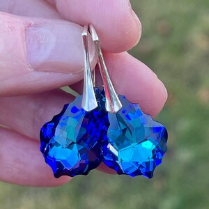 May include: A pair of faceted, deep blue crystal earrings with silver-colored hooks. The earrings are held up against a blurred green background, showcasing their vibrant color and intricate cut.