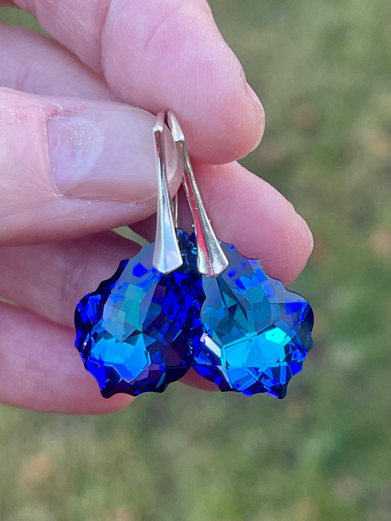 May include: A pair of faceted, deep blue crystal earrings with silver-colored hooks. The earrings are held up against a blurred green background, showcasing their vibrant color and intricate cut.