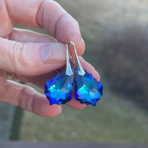 May include: A pair of blue crystal drop earrings with silver-colored hooks. The earrings have a faceted, teardrop shape, reflecting light with shades of blue and teal. The jewelry is held up against a blurred outdoor background.