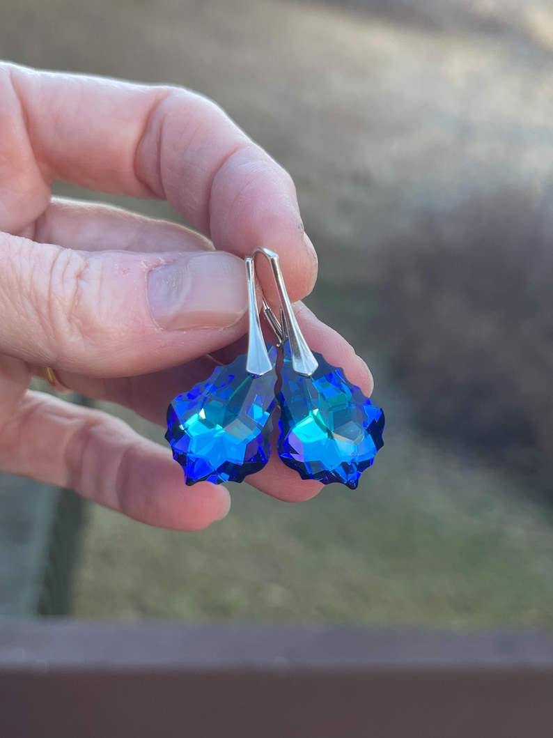 May include: A pair of blue crystal drop earrings with silver-colored hooks. The earrings have a faceted, teardrop shape, reflecting light with shades of blue and teal. The jewelry is held up against a blurred outdoor background.