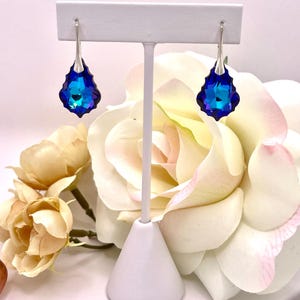 May include: A pair of drop earrings with a silver-tone hook and a large, faceted, teardrop-shaped blue crystal. The crystals have a gradient of blue hues, from deep indigo to bright turquoise. The earrings are displayed on a white jewelry stand, with a backdrop of cream and pink roses.