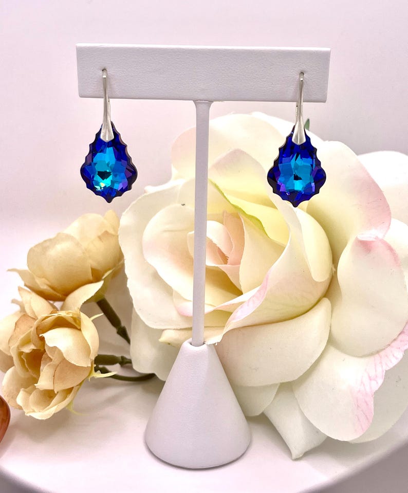May include: A pair of drop earrings with a silver-tone hook and a large, faceted, teardrop-shaped blue crystal. The crystals have a gradient of blue hues, from deep indigo to bright turquoise. The earrings are displayed on a white jewelry stand, with a backdrop of cream and pink roses.