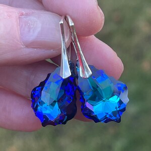 May include: A pair of faceted, teardrop-shaped earrings. The earrings feature a vibrant blue color with hints of green and silver-tone metal hooks. The earrings are held up against a blurred green background.
