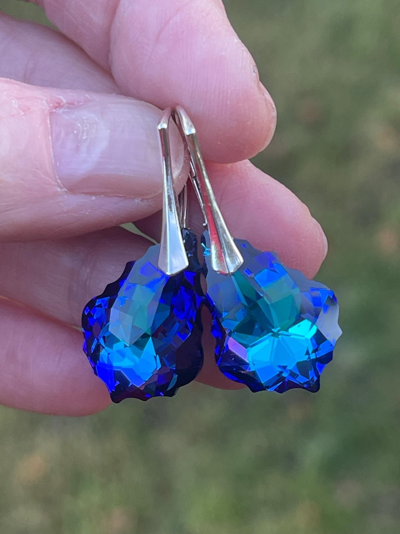 May include: A pair of faceted, teardrop-shaped earrings. The earrings feature a vibrant blue color with hints of green and silver-tone metal hooks. The earrings are held up against a blurred green background.