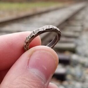 May include: A silver ring with a textured band, held in a hand against a blurred background of railroad tracks.