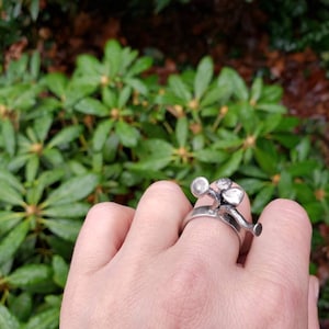 Könnte beinhalten: Ein silberner Ring mit einem dreidimensionalen Design einer Blume mit drei Blütenblättern und einem Stiel. Der Ring befindet sich an einem Finger.