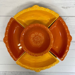 May include: A four-piece ceramic serving dish set in shades of orange and yellow. The set features a central bowl and three smaller bowls that fit around the central bowl. The bowls have a floral design.